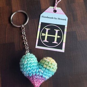 Puede incluir: Un llavero de coraz&oacute;n de crochet arco&iacute;ris con un anillo y una cadena plateados. El llavero tiene una etiqueta que dice "Handmade by Hennek" con una H en blanco y negro con una flecha que lo atraviesa.