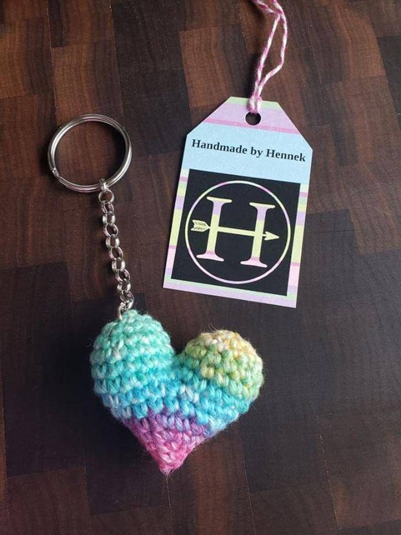 Puede incluir: Un llavero de coraz&oacute;n de crochet arco&iacute;ris con un anillo y una cadena plateados. El llavero tiene una etiqueta que dice "Handmade by Hennek" con una H en blanco y negro con una flecha que lo atraviesa.
