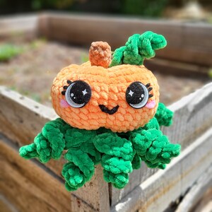 May include: A crocheted pumpkin plush toy with a green stem and leaves. The pumpkin has a smiling face with large black eyes and pink cheeks.