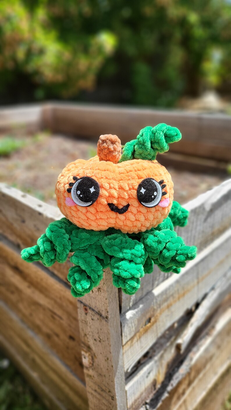 May include: A crocheted pumpkin plush toy with a green stem and leaves. The pumpkin has a smiling face with large black eyes and pink cheeks.
