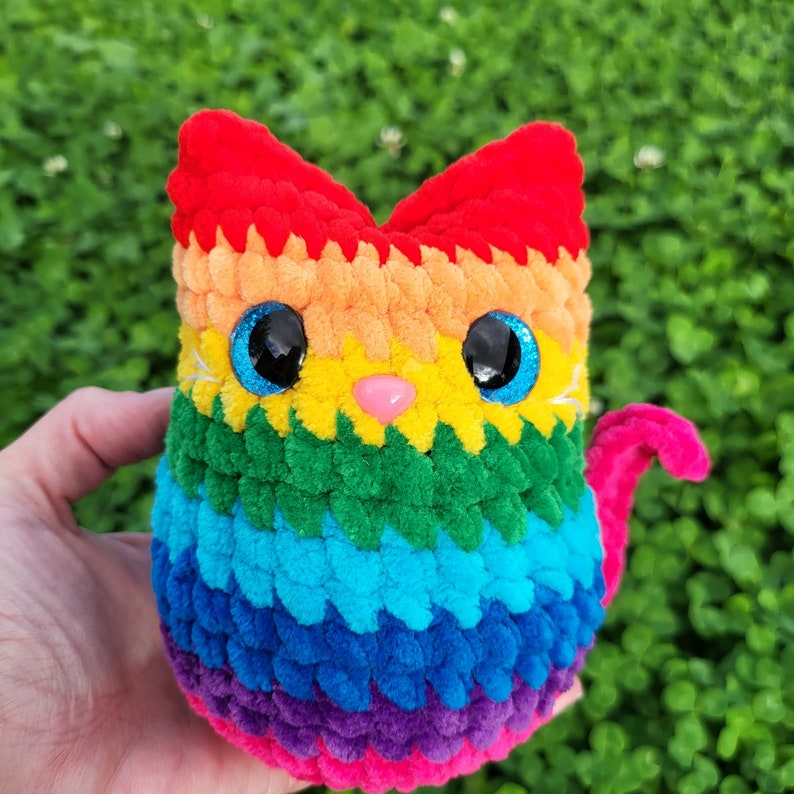 May include: A rainbow crocheted cat toy with blue eyes and a pink nose. The cat is made of yarn in shades of red, orange, yellow, green, blue, purple, and pink.