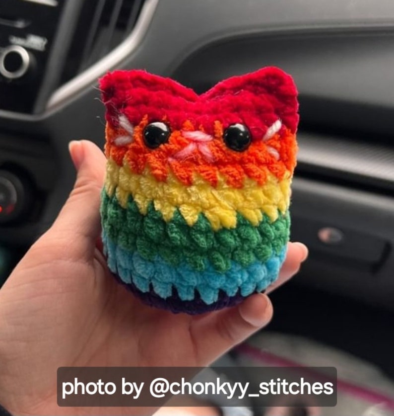 May include: A rainbow crocheted cat plush toy with black eyes and a white nose. The toy is being held in a person's hand. Photo by @chonkyy_stitches