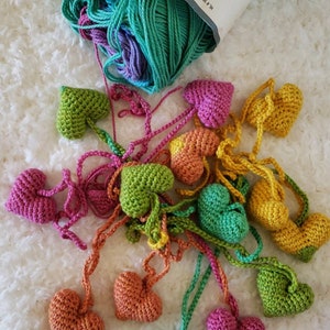 Puede incluir: Una guirnalda de corazones tejidos a crochet en varios colores, incluyendo verde, rosa, naranja y amarillo. Los corazones est&aacute;n unidos con hilo y cuelgan de una superficie blanca. El texto "SWOONING HEART GARLAND" est&aacute; impreso debajo de los corazones.