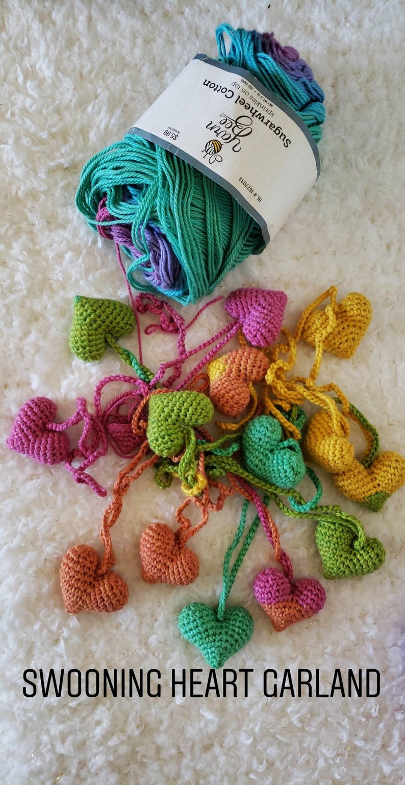 Puede incluir: Una guirnalda de corazones tejidos a crochet en varios colores, incluyendo verde, rosa, naranja y amarillo. Los corazones est&aacute;n unidos con hilo y cuelgan de una superficie blanca. El texto "SWOONING HEART GARLAND" est&aacute; impreso debajo de los corazones.