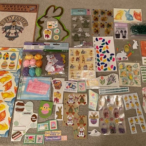 May include: A collection of Easter-themed craft supplies, including stickers, decorative eggs, and paper cutouts. The items feature bunnies, eggs, and spring-themed text. Colors include pastels, greens, and yellows. The phrase "Happy Easter" is visible on a few items.