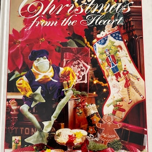 May include: A book cover featuring a festive Christmas scene with a frog wearing a blue and yellow outfit, a stocking with a nutcracker design, and a gingerbread person. The book title is "Christmas from the Heart" and the subtitle is "Holiday Magic".