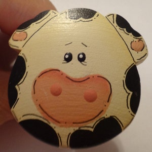 May include: A wooden cow-shaped knob with black and white spots, a pink nose, and black eyes. The knob is painted with a whimsical style.