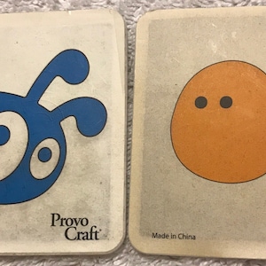 May include: Two square coasters with cartoon designs. One coaster features a blue bug-like creature, the other an orange fish. Both have a light beige background and the text "Provo Craft" and "Made in China".