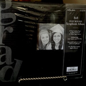 May include: Black 8x8 post-bound scrapbook album with a photo of two women in graduation caps. The album has 20 top-loading pages, 10 sheet protectors, and 10 white paper inserts. The album is archival safe.