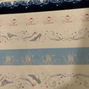 May include: Four strips of decorative washi tape featuring a Cinderella theme. The top strip is a dark blue background with white and silver stars and a white and silver design. The second strip is white with a pink crown and the word "Cinderella" in pink. The third strip is white with a blue castle silhouette and silver stars. The fourth strip is white with a silver glass slipper and a silver swirl design.