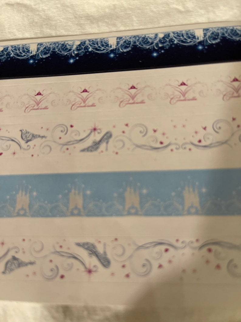 May include: Four strips of decorative washi tape featuring a Cinderella theme. The top strip is a dark blue background with white and silver stars and a white and silver design. The second strip is white with a pink crown and the word "Cinderella" in pink. The third strip is white with a blue castle silhouette and silver stars. The fourth strip is white with a silver glass slipper and a silver swirl design.