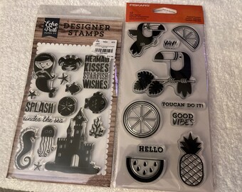 May include: Two sets of clear rubber stamps, one with a mermaid and ocean theme and the other with a toucan and tropical fruit theme. The mermaid set includes stamps of a mermaid, a starfish, a castle, a seahorse, a jellyfish, and the words "Mermaid Kisses, Starfish Wishes, Splash Under the Sea". The toucan set includes stamps of a toucan, a pineapple, a watermelon, a lemon, and the words "Toucan Do It!, Good Vibes, Yay!, Hello".