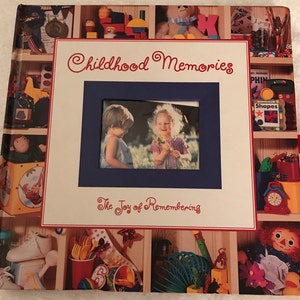 May include: A white book with a red and blue border. The cover features a photo of two children in a field, a title that reads "Childhood Memories", and the text "The Joy of Remembering". The cover is decorated with images of toys and other childhood items.