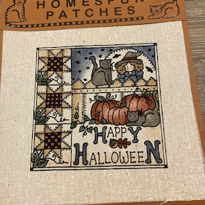 May include: Homespun patches with a Halloween theme. The image features a square patch with a painted design of pumpkins, a witch, and cats. The words "HAPPY HALLOWEEN" are written in blue. The patch is on a tan background.