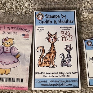May include: A collection of cat-themed stamps. The stamps include images of cartoon cats, phrases like "I Love You," and "May paw prints be forever on your heart." The stamps are packaged in clear plastic and labeled with the brand names "Sweet Impressions Stamps" and "Stamps by Judith & Heather."