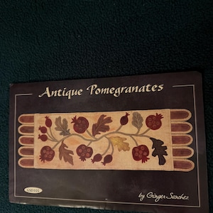 May include: A dark-colored package with the text "Antique Pomegranates" and a decorative pomegranate design. The design features pomegranates, leaves, and stems in shades of red, brown, and gold. The package is labeled "GSD105" and "by Ginger Sanchez."