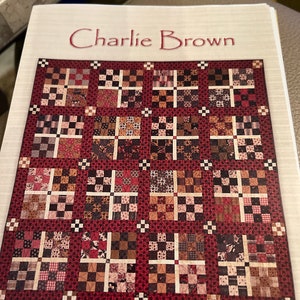 May include: A quilt pattern titled "Charlie Brown" featuring a red background with a patchwork design of various fabrics in shades of brown, black, white, and red.