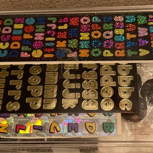 May include: A clear plastic storage box filled with various sticker sheets. The sheets contain colorful alphabet letters, numbers, and other symbols. The box also includes a small, round, black sticker.