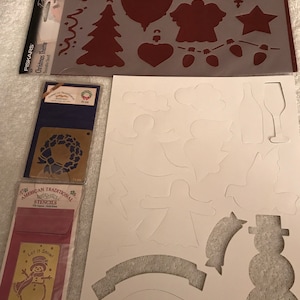 May include: A set of Christmas stencils featuring various holiday designs, including a snowman, angels, a wreath, a tree, a star, and a bell. The stencils are made of plastic and are designed for use with paint or other crafting materials.