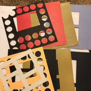 May include: A collection of colorful mat boards in various shapes and sizes. The mats are in shades of red, black, gold, peach, and blue. Some mats have circular cutouts, while others have rectangular openings. The mats are likely for framing artwork or photographs.
