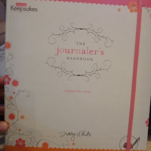 May include: A hardcover journal titled "The Journaler's Handbook" with a pink and orange border. The cover features floral designs and the text "everyone has a story". A pink elastic band is visible on the right side.
