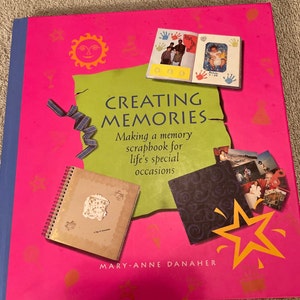 May include: A pink book with a green and black scrapbook on the cover. The book's title is "Creating Memories: Making a memory scrapbook for life's special occasions" by Mary-Anne Danagher.