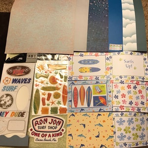 May include: A set of colorful scrapbook paper featuring a beach theme with surfboards, waves, and flowers. The paper includes the text "Surfs Up!" and "Ron Jon Surf Shop, One of a Kind, Cocoa Beach, Fla."