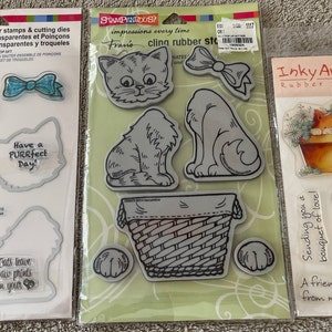 May include: Three packages of cat-themed rubber stamps and cutting dies. The stamps feature various cat illustrations, bows, and phrases like "Love Mew" and "Have a PURRfect Day!" The stamps are in clear plastic packaging.