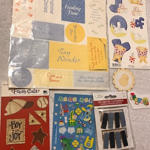 May include: A collection of baby-themed scrapbook stickers and embellishments. Includes stickers with phrases like "Tiny Wonder" and "Baby Boy", along with decorative clothespins and die-cut shapes. Colors include blue, yellow, and red.