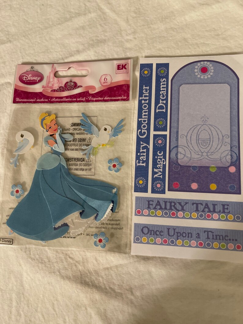 May include: A set of six dimensional Cinderella stickers featuring Cinderella, a carriage, and birds. The stickers are on a clear plastic sheet with a pink and white background. The text "Fairy Godmother Magic Dreams" and "Fairy Tale Once Upon a Time..." is printed on the sheet.