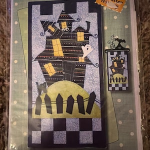 May include: A Halloween-themed fabric panel featuring a haunted house design. The panel, measuring 6 x 12 inches, showcases a black house with yellow windows, a ghost, and a fence. The background is a swirl pattern with a green and black checkered border.