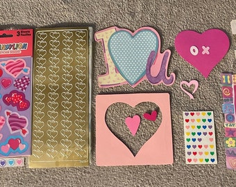 May include: A set of Valentine's Day craft supplies featuring stickers, paper, and cutouts. The set includes a variety of hearts, flowers, and other designs in pink, red, and white. The set also includes a small book with a pink ribbon.