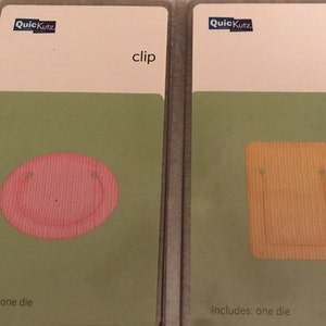 May include: Two QuickKutz clip dies in clear plastic packaging. One die is a pink circle with a smiley face design, and the other is a tan rectangle. The packaging has a white top with the brand name and the word "clip".