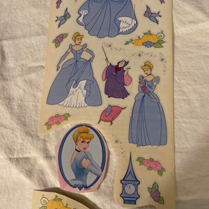 May include: A sheet of stickers featuring Cinderella in various poses, including her iconic blue ball gown, a pink dress, and a portrait. The sheet also includes a sticker of the evil stepmother, a pumpkin, a clock, and birds.
