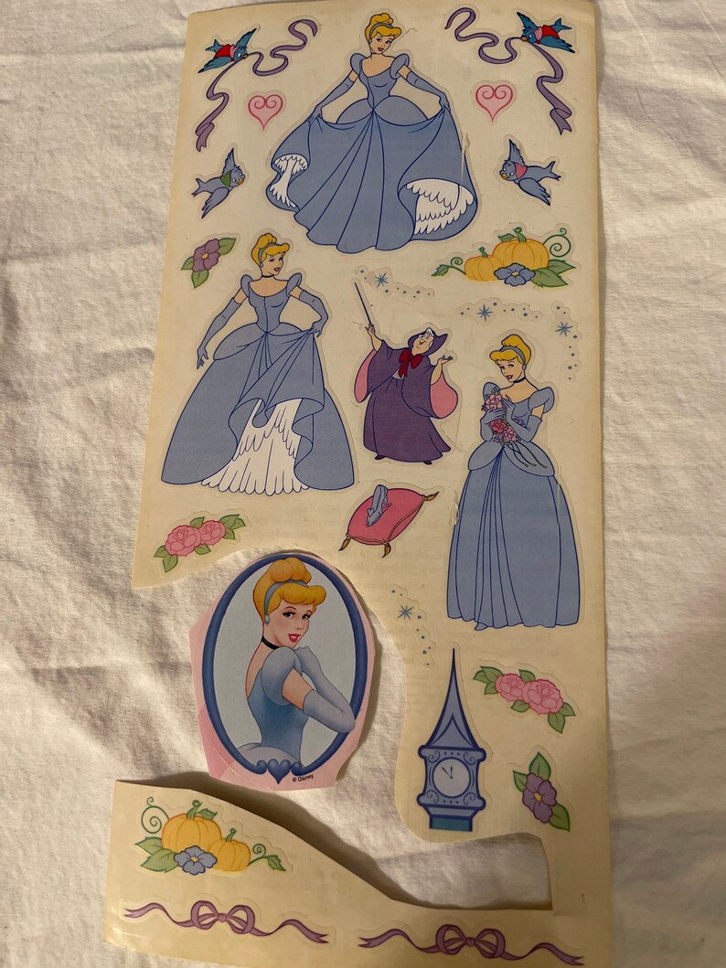 May include: A sheet of stickers featuring Cinderella in various poses, including her iconic blue ball gown, a pink dress, and a portrait. The sheet also includes a sticker of the evil stepmother, a pumpkin, a clock, and birds.