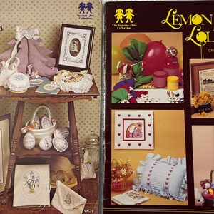 May include: Two cross-stitch pattern books, "Sweet Memories" and "Lemon Drops and Lollipops". The covers feature colorful illustrations of cross-stitch designs, including a tree, a heart, a rainbow, and a house.