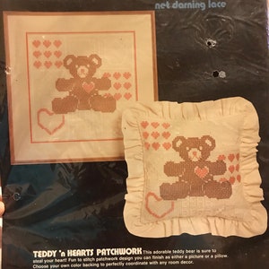 May include: A cross-stitch kit featuring a teddy bear surrounded by hearts. The kit includes 6 mesh net fabric, perle cotton thread, a needle, and easy instructions. The finished pillow size is 12 inches by 12 inches, and the frame size is 16 inches by 16 inches.