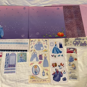 May include: A set of Cinderella themed scrapbooking supplies, including paper, stickers, and embellishments. The paper is purple and pink with a star pattern. The stickers feature Cinderella, the fairy godmother, and other characters from the movie. The embellishments include a ruler, a die cut, and a sticker sheet.