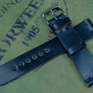 May include: A navy blue leather watch strap with a silver buckle. The strap has visible stitching in a contrasting blue thread. The background is a textured green surface with the text "HORWEEN 1905".