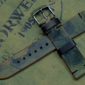 Op de afbeelding: Een groen en bruin leren horlogebandje met zwarte stiksels en een zilveren gesp.