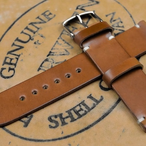 May include: A brown leather watch strap with a silver buckle and white stitching. The strap is made of high-quality leather and is designed to fit most standard watch sizes. The background features a leather surface with the words "GENUINE SHELL" printed on it.