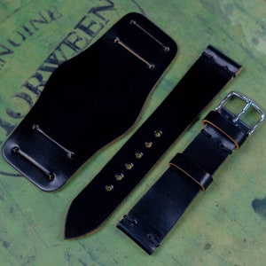 Puede incluir: Juego de correas de reloj de cuero negro, que incluye una pulsera ancha, una correa larga con múltiples agujeros y una correa más corta con una hebilla plateada. Se muestra sobre una superficie texturizada verde y marrón.