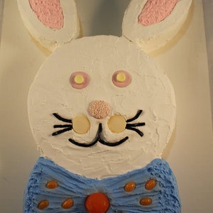 May include: A white bunny cake with pink ears and a blue bow tie decorated with brown candy. The cake is on a white surface.