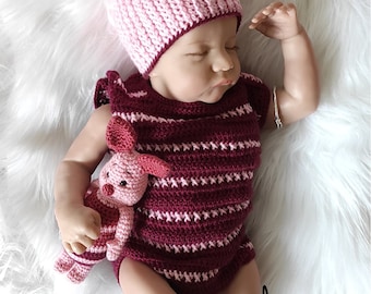 May include: A baby wearing a pink and burgundy striped crocheted onesie and a pink and burgundy crocheted hat with bunny ears. The baby is holding a pink crocheted pig toy. The baby is lying on a white fluffy surface.