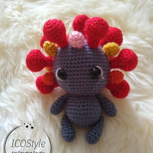 Könnte beinhalten: Ein gehäkeltes Axolotl-Plüschtier mit grauem Körper, roten und orangen Kiemen und schwarzen Augen. Das Spielzeug sitzt auf einer weißen, flauschigen Oberfläche.