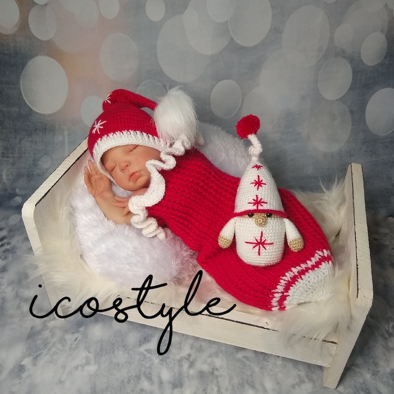 Pode incluir: Um conjunto de beb&eacute; de crochet vermelho e branco com um boneco gnomo a condizer. O conjunto tem uma bainha de babados brancos e um pompom branco no chap&eacute;u. O boneco gnomo tem uma barba branca e um chap&eacute;u vermelho com estrelas brancas.