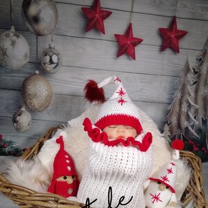 Pode incluir: Um saco de dormir para beb&ecirc; de croch&ecirc; branco com detalhes em vermelho e um chap&eacute;u de croch&ecirc; combinando. O beb&ecirc; est&aacute; cercado por dois enfeites de gnomo de croch&ecirc; com chap&eacute;us vermelhos e barbas brancas. O fundo &eacute; uma parede de madeira r&uacute;stica com estrelas vermelhas penduradas no teto.