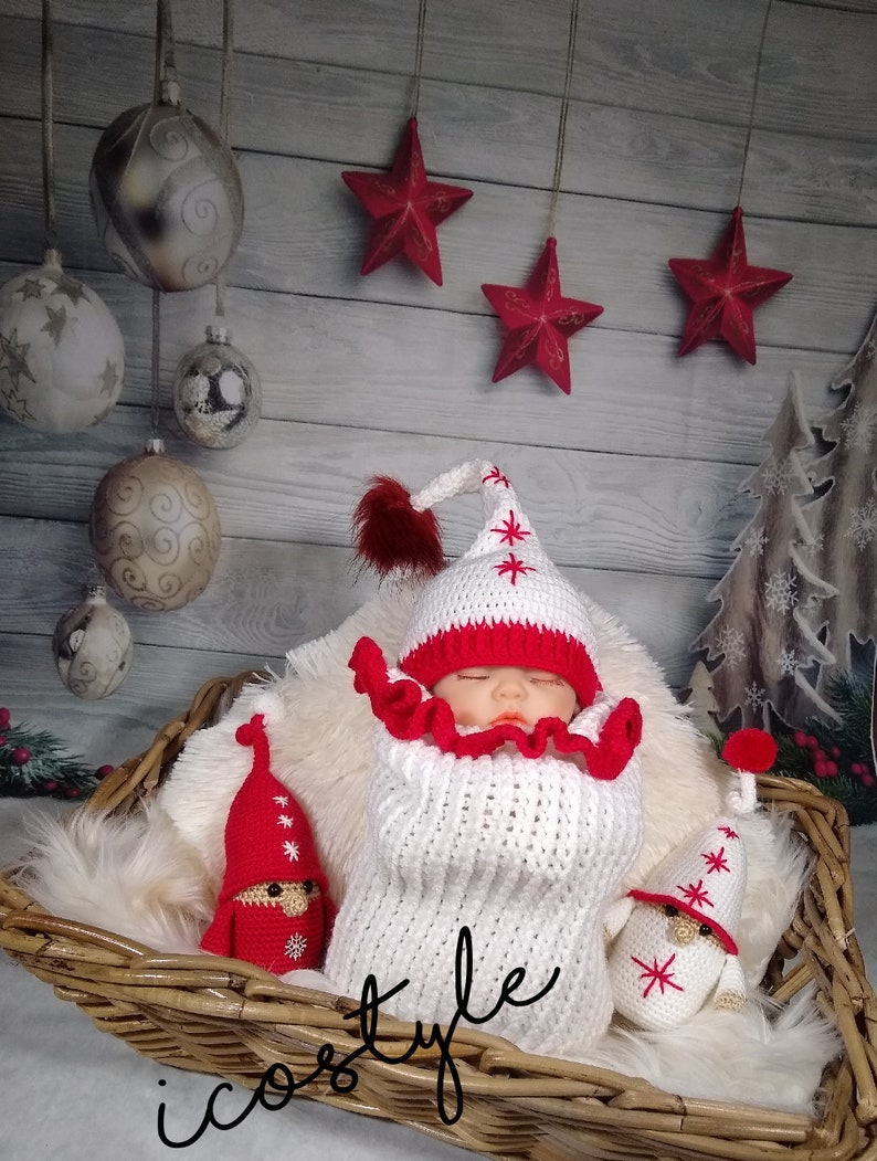 Pode incluir: Um saco de dormir para beb&ecirc; de croch&ecirc; branco com detalhes em vermelho e um chap&eacute;u de croch&ecirc; combinando. O beb&ecirc; est&aacute; cercado por dois enfeites de gnomo de croch&ecirc; com chap&eacute;us vermelhos e barbas brancas. O fundo &eacute; uma parede de madeira r&uacute;stica com estrelas vermelhas penduradas no teto.