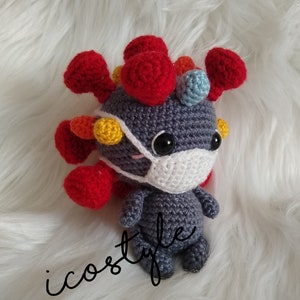 May include: A gray crocheted plush toy with a white face mask. The toy has red, yellow, and blue crocheted spikes on its head.