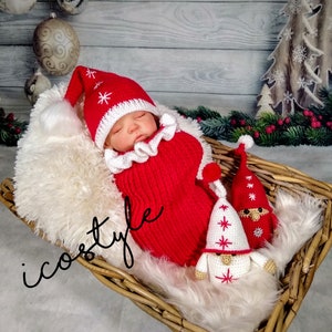 Pode incluir: Um chap&eacute;u de Papai Noel de croch&ecirc; vermelho e branco e um conjunto combinando para um beb&ecirc;, com dois gnomos de croch&ecirc; branco com detalhes vermelhos. O beb&ecirc; est&aacute; dormindo em uma cesta de vime forrada com pele branca.